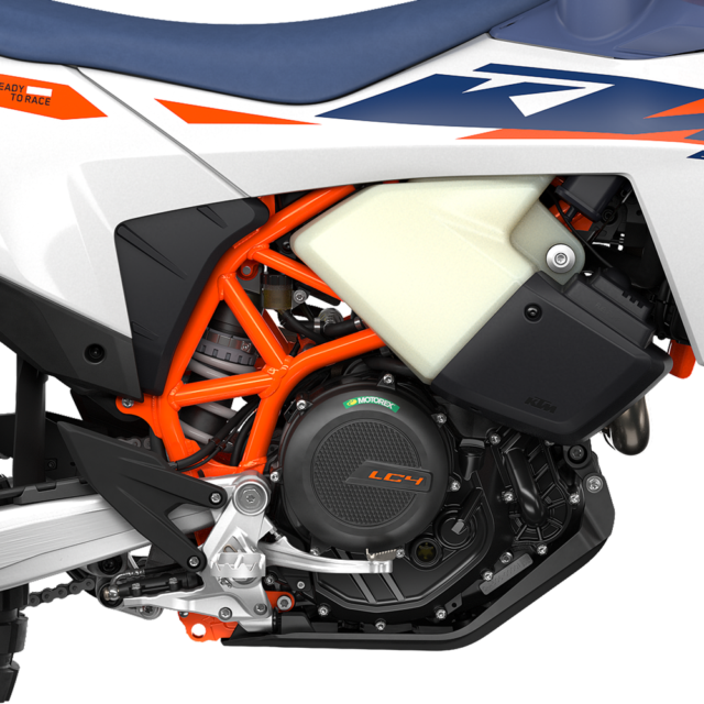 Close-up of a 2027 KTM 690 Rally motorcycle engine with a bright orange frame, black casing marked LC4, and white and blue body panels. Parts of the gearbox, exhaust, and protective covers are visible.