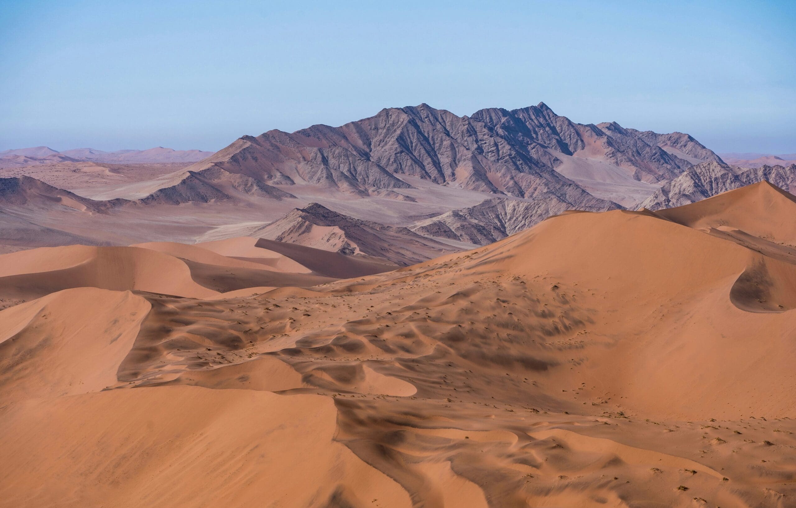 Expansive desert landscape with rolling orange sand dunes in the foreground and jagged rocky mountains in the background under a clear blue sky—a scene straight from an Overland News feature.