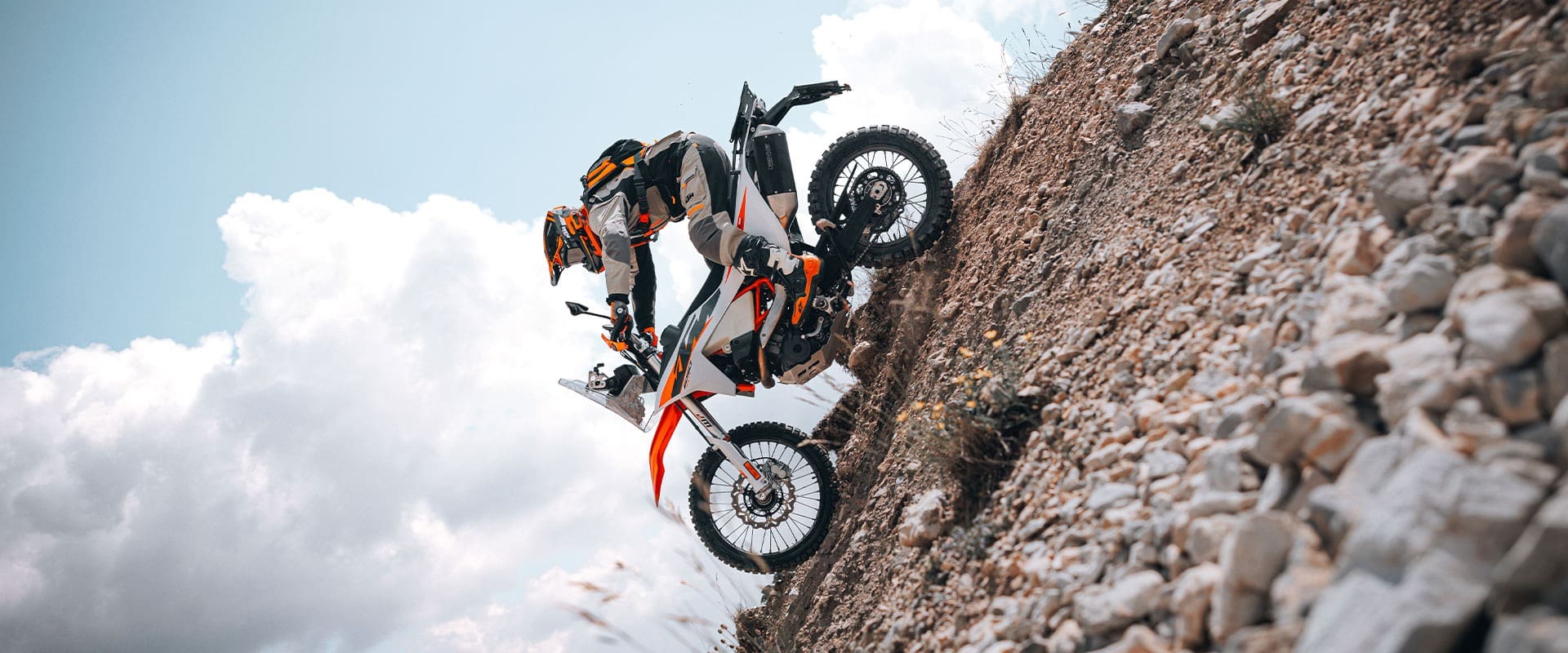 A motorcyclist in full gear rides a 2027 KTM 690 Rally dirt bike up a steep, rocky hill against a cloudy sky, demonstrating off-road skills and control.
