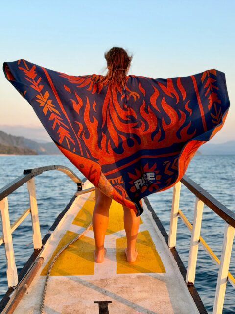A person stands barefoot on a dock over water, facing away, holding a large blue and orange towel with a flame-like design spread wide, with mountains visible in the background—a peaceful scene that could easily grace the cover of Overland News.