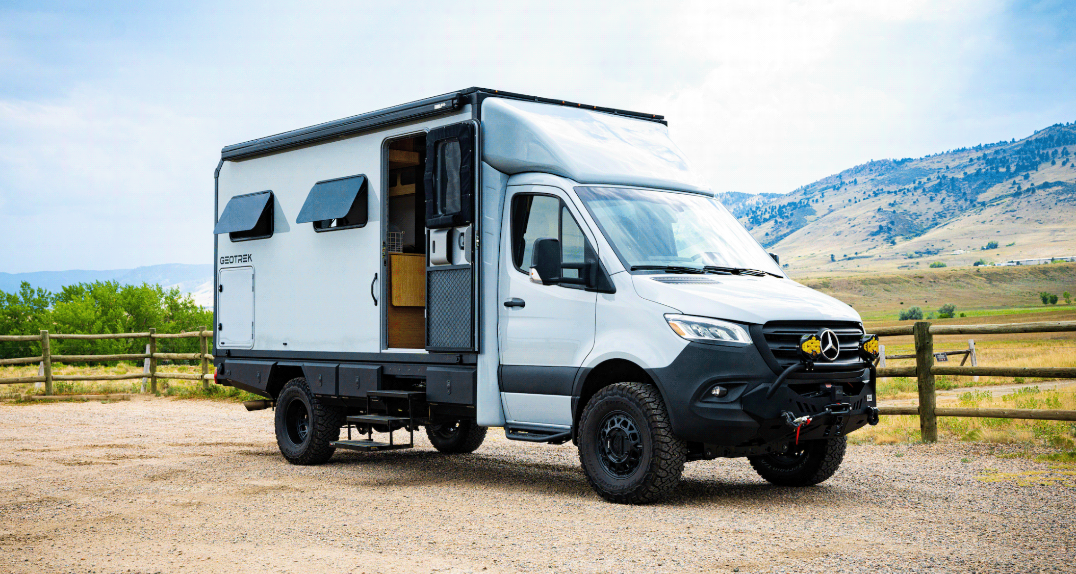 A white camper van with off-road tires is parked on gravel near a wooden fence, with mountains and a partly cloudy sky in the background. The van’s side door is open, revealing part of the interior—perfect for anyone seeking inspiration from Overland News.