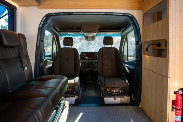 Interior view of a camper van featured in Overland News, showing two front seats, a sofa on the left, wooden cabinetry on the right, and a fire extinguisher mounted to the wall. The windshield reveals a rocky outdoor landscape.