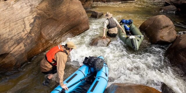 Two people wearing hats and life vests guide inflatable kayaks through shallow, rocky rapids in a narrow stream, surrounded by large rocks and flowing water—a true Overland News moment for adventurous explorers.