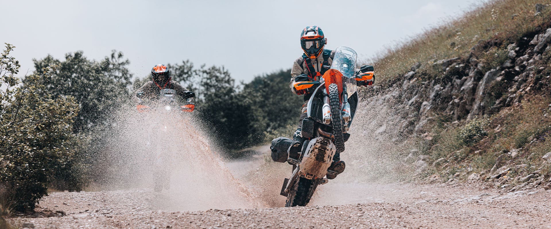Two motorcyclists ride off-road on a gravel trail aboard their 2027 KTM 690 Rally bikes. The rider in front is lifting the front wheel in a wheelie, while the second rider follows, kicking up water and dirt as they speed through a wet section of the path.