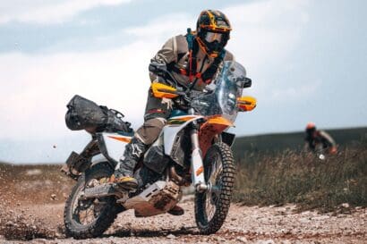 A person wearing protective gear rides a 2027 KTM 690 Rally on a rocky trail, kicking up dust and gravel, with another rider visible in the background under a cloudy sky.