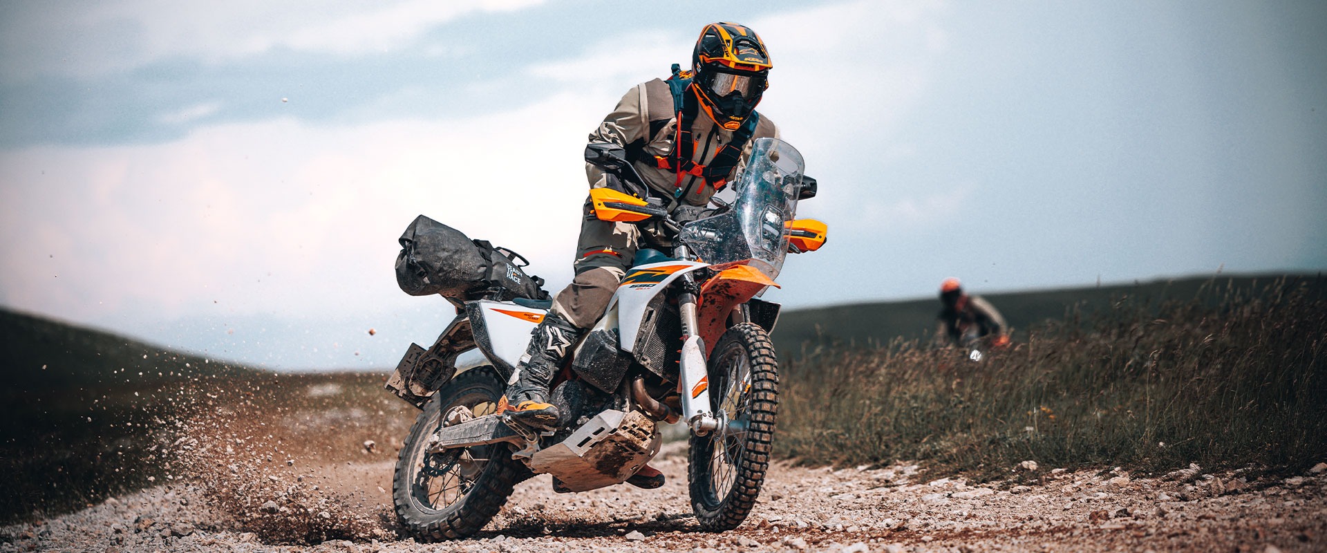A person wearing protective gear rides a 2027 KTM 690 Rally on a rocky trail, kicking up dust and gravel, with another rider visible in the background under a cloudy sky.