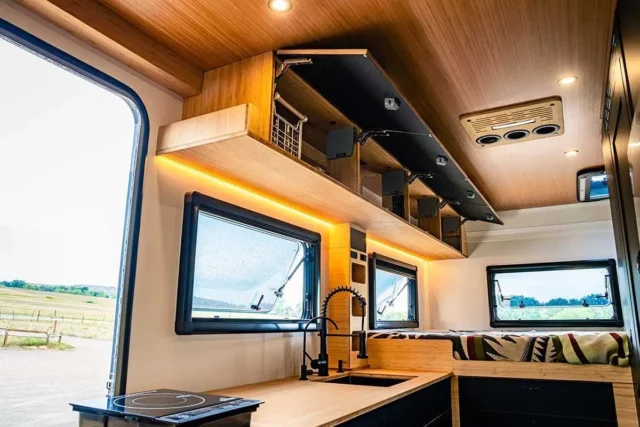Modern camper van interior with open storage cabinets, wood paneling, windows, a sink, stove top, and cushioned seating; daylight streams in, revealing a scenic outdoor view—perfect for Overland News enthusiasts seeking inspiration for their next adventure.