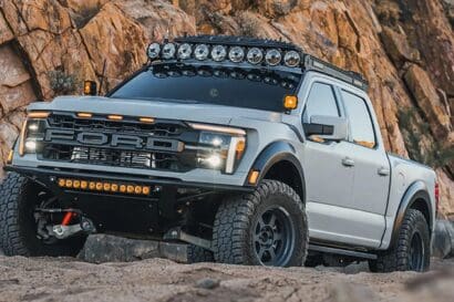 A silver Ford pickup truck with off-road tires, roof-mounted lights, and a rugged front bumper is parked on sandy terrain near a rocky cliff, making it an eye-catching sight for any Overland News enthusiast.