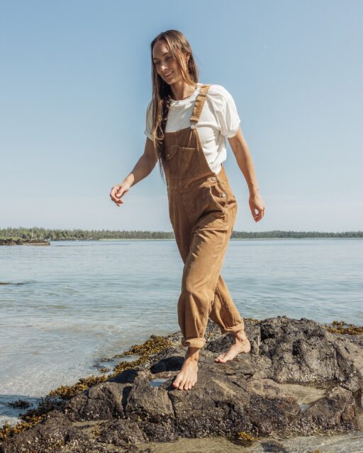 A person wearing a white shirt and brown overalls stands barefoot on a rocky shore, soaking in the beauty of nature with the ocean and trees in the background under a clear blue sky—a perfect scene for an Overland News feature.