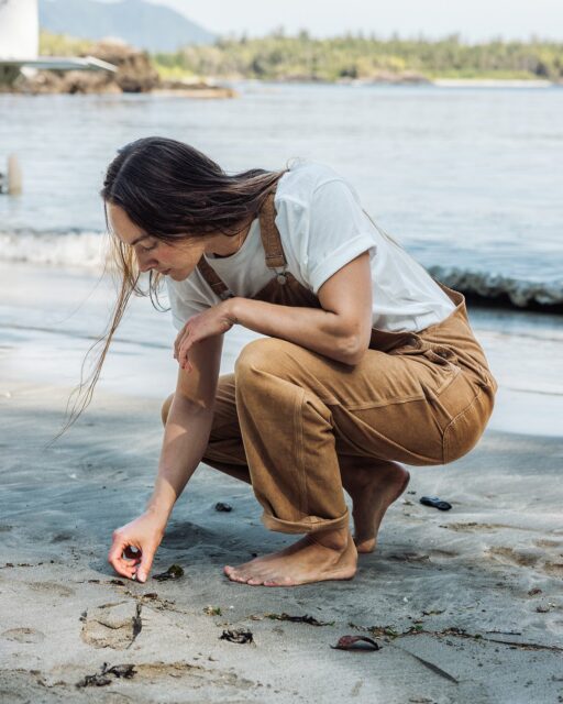 A person in brown overalls and a white shirt squats barefoot on a sandy beach, using a finger to draw or touch something in the sand. Water and trees are visible in the background, creating a peaceful scene reminiscent of an Overland News feature.