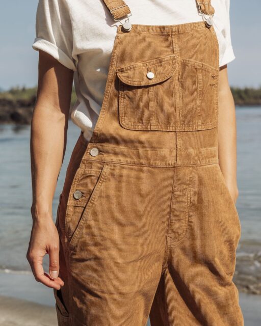 A person wearing a white T-shirt and light brown corduroy overalls stands near water with a shoreline and trees visible in the blurred background, showcasing relaxed outdoor style perfect for an Overland News feature.