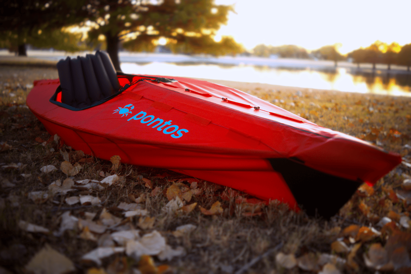 A bright red kayak labeled pontos sits on dry grass near a calm lake at sunset, surrounded by fallen leaves and trees—ready for its next adventure, as featured in Overland News.
