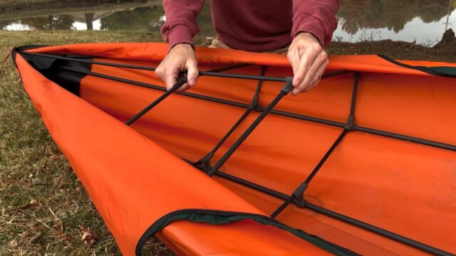 A person in a red sweatshirt assembles the black frame of an orange folding kayak on grass near a body of water, capturing an adventurous moment perfect for Overland News.