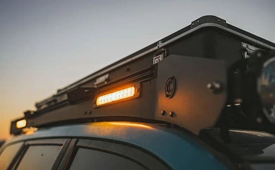 Close-up of a vehicle roof rack with an illuminated amber light bar, mounted on top of a blue car, taken at sunset with a clear sky in the background—perfect for sharing the latest Overland News.