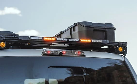 The image shows the rear roof of a vehicle with a rooftop cargo box, roof rack, and illuminated amber hazard lights mounted along the rack—perfectly capturing an Overland News-worthy setup for adventure enthusiasts.