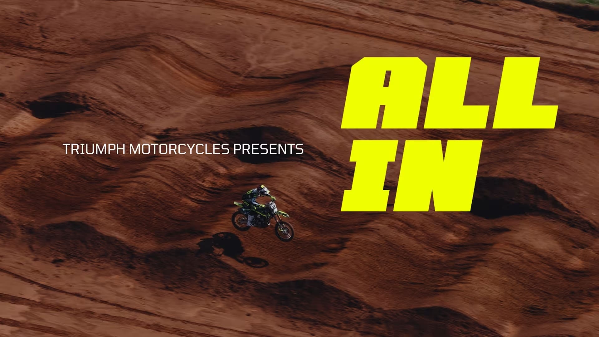 A motocross rider jumps on a dirt track. Large yellow text says ALL IN, and smaller white text reads TRIUMPH MOTORCYCLES PRESENTS, featured by Overland News.