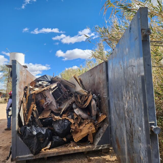 A metal dumpster filled with black trash bags, cardboard, and assorted debris sits outdoors beneath a blue sky with clouds and surrounding tall grass—an everyday scene captured by Overland News.