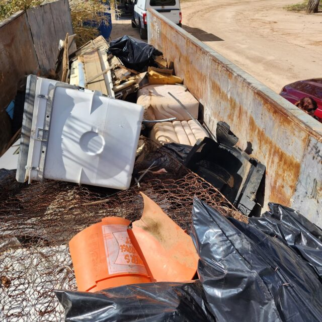 A dumpster filled with discarded items, including a mattress spring, a white appliance, black trash bags, an orange container, and various debris, sits outdoors on a sunny day—an all-too-familiar sight captured recently by Overland News.