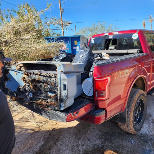 A red pickup truck filled with burned and damaged household items and trash bags, its tailgate blackened and melted, parked on a dirt surface near bushes and a blue dumpster, creates a scene straight out of Overland News.