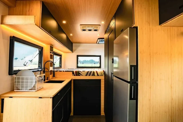 Modern camper van interior with wood cabinetry, black accents, a kitchen area with a sink and fridge, and a small bed in the back near windows. Natural light illuminates the space, creating a cozy atmosphere—perfect for catching up on Overland News or planning your next adventure.