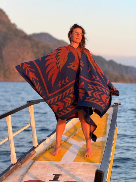A person wearing sunglasses and a blue-and-red poncho stands barefoot on the bow of a boat, with mountains and water in the background during sunset—a peaceful moment captured for Overland News.
