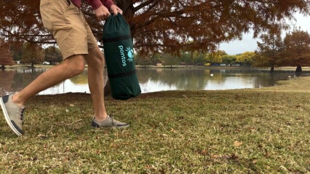 A person in shorts and slip-on shoes carries a green bag labeled Sonos while walking on grass near a lake, with a large tree and calm water in the background—capturing an idyllic moment worthy of Overland News.