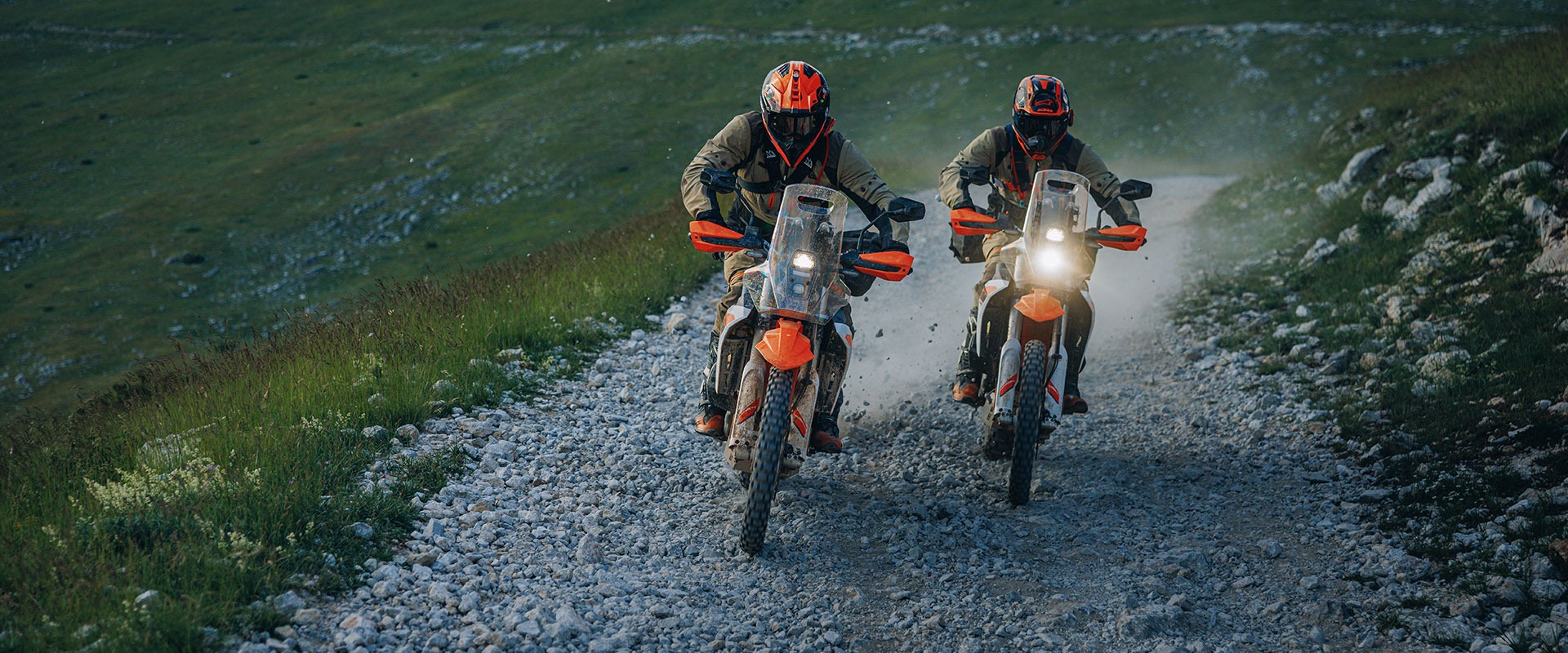 Two people in helmets and gear ride 2027 KTM 690 Rally orange dirt bikes side by side on a rocky, gravel road surrounded by grassy hills. Dust rises behind them as they travel.