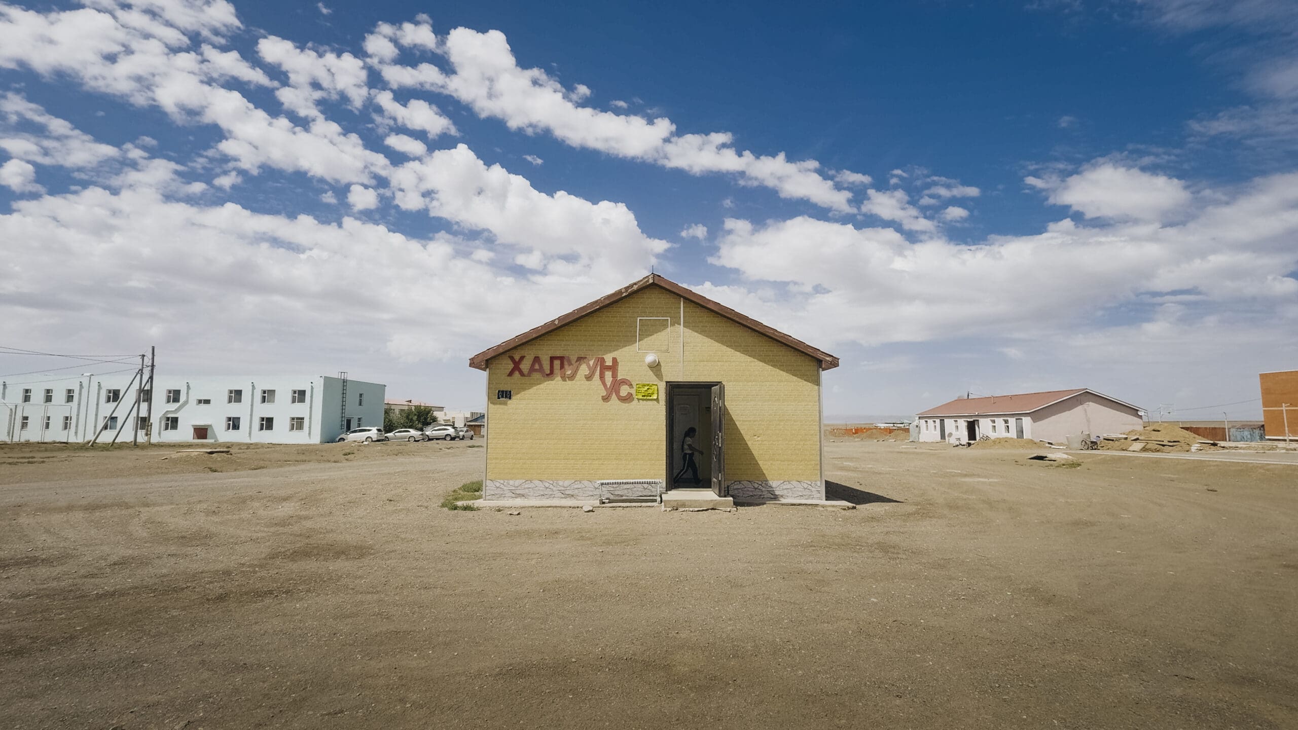 A small yellow building with text on its wall stands alone on a dirt lot under a partly cloudy sky, embodying the remote charm often encountered while overlanding Mongolia, with a few other buildings visible in the background.