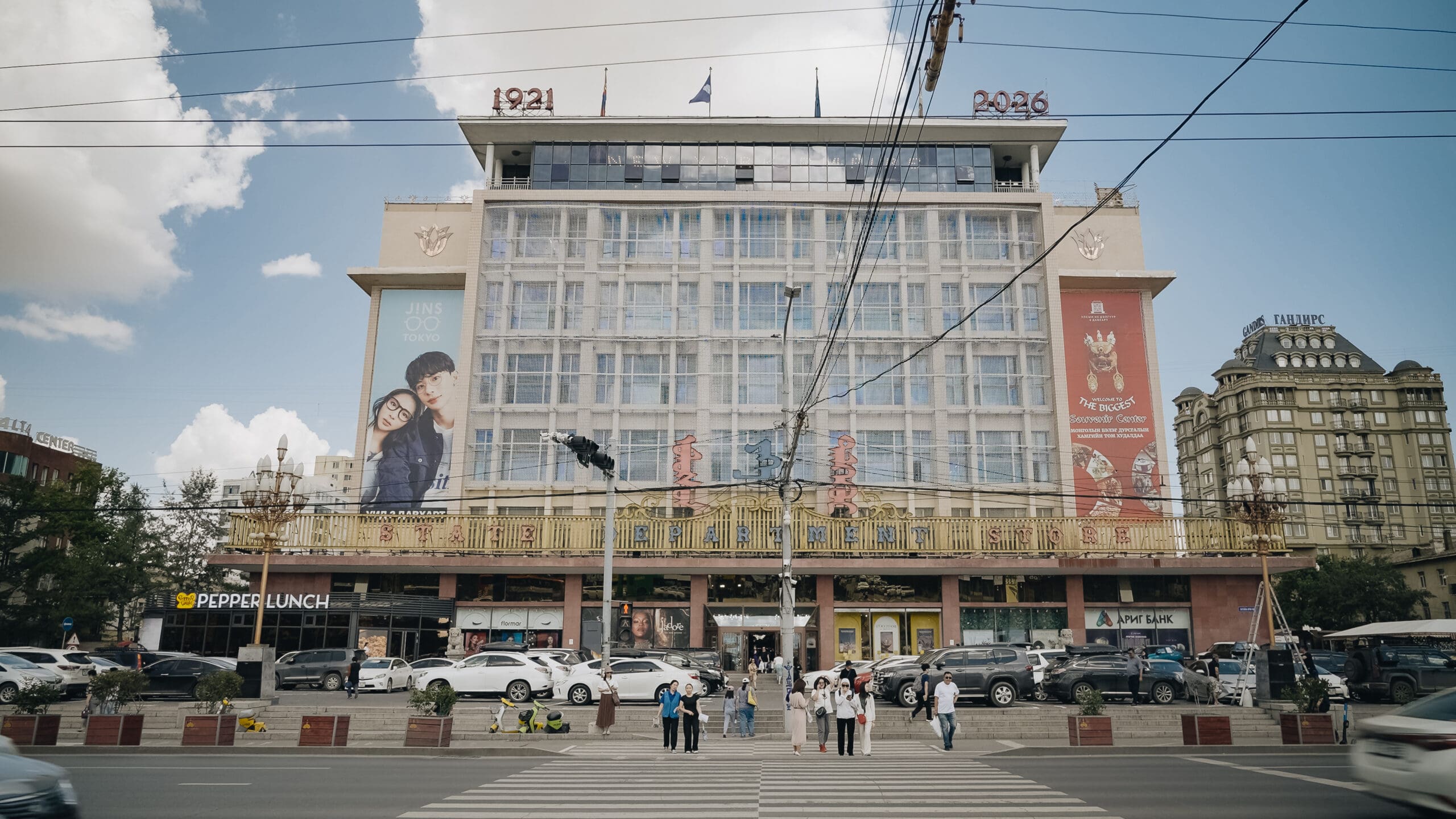A large building with glass windows, banners, and people crossing a street in front. Cars are parked along the roadside. Flags and numbers 1921 and 2026 are displayed on the roof, while visitors browse nearby stalls offering souvenirs.