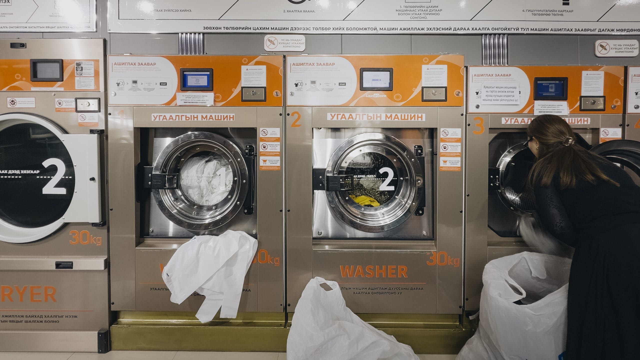A woman loads laundry into one of several large washing machines at a laundromat—a much-needed stop after days of overlanding Mongolia. Each machine is labeled WASHER and 30kg in English and another language. White laundry bags are in the foreground.