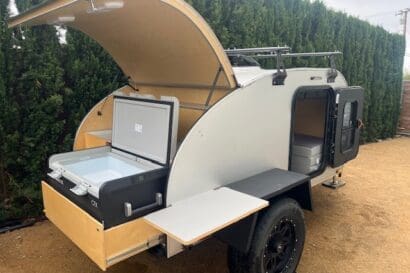 A compact 2021 teardrop trailer with an open rear hatch reveals a slide-out cooler, wooden shelves, and a side door open to a small interior. The RAD trailer is parked on a gravel surface next to tall greenery.