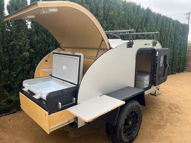 A compact 2021 teardrop trailer with an open rear hatch reveals a slide-out cooler, wooden shelves, and a side door open to a small interior. The RAD trailer is parked on a gravel surface next to tall greenery.