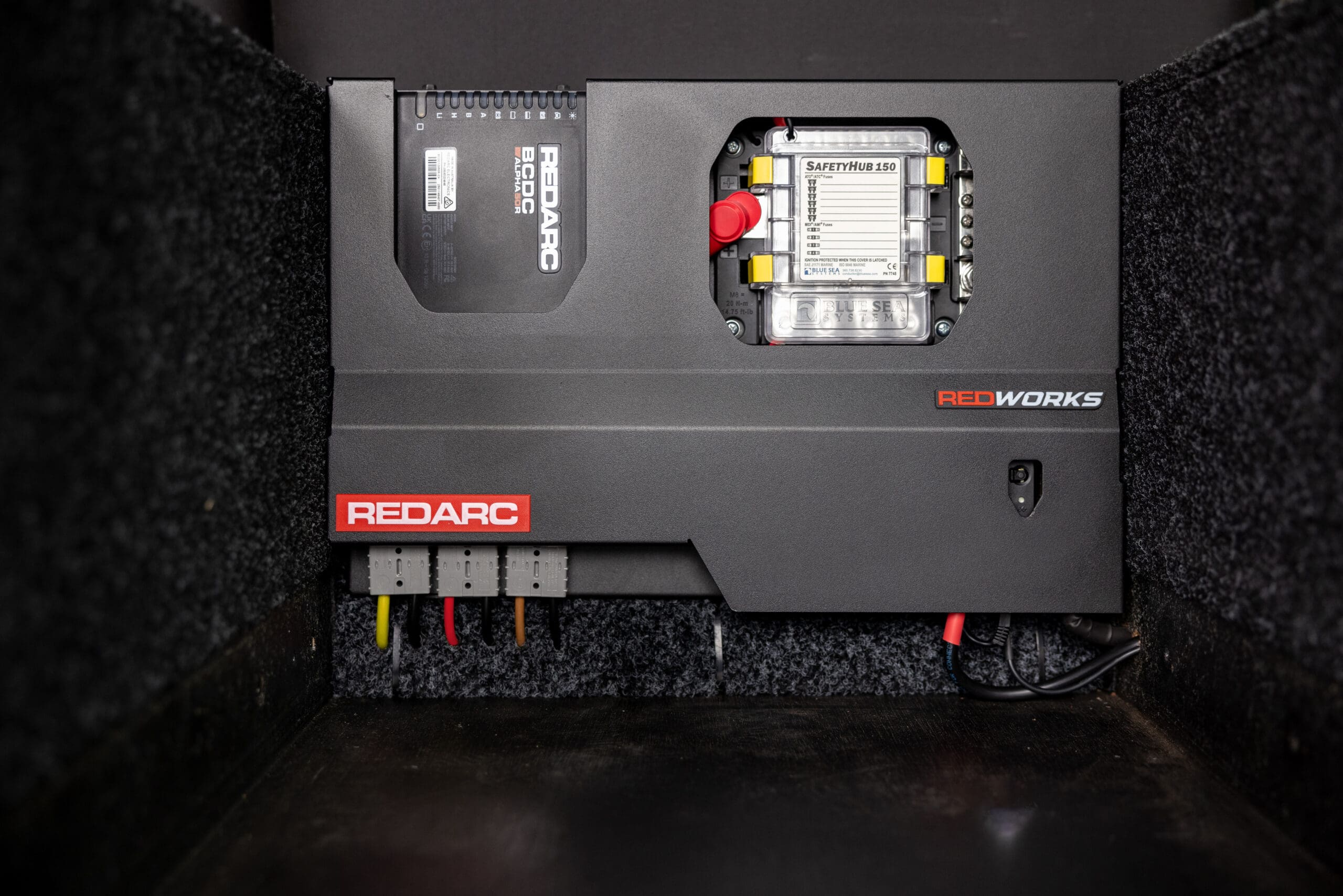 A Redarc power management unit installed in a compartment, featuring a BCDC charger, a SafetyHub 150 fuse block, and electrical connections for weekly overland news updates.