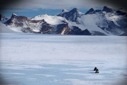 Two vehicles from Expeditions 7 travel across the vast, snow-covered landscape of the White Desert toward distant snow-capped mountains.