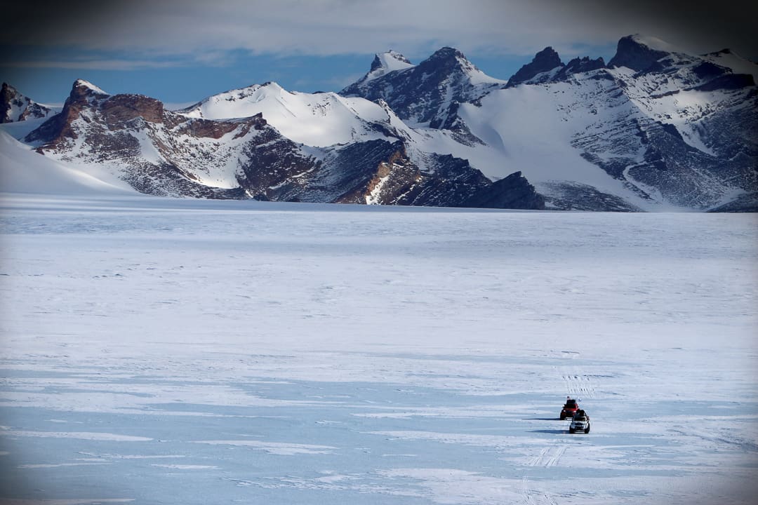 Two vehicles from Expeditions 7 travel across the vast, snow-covered landscape of the White Desert toward distant snow-capped mountains.