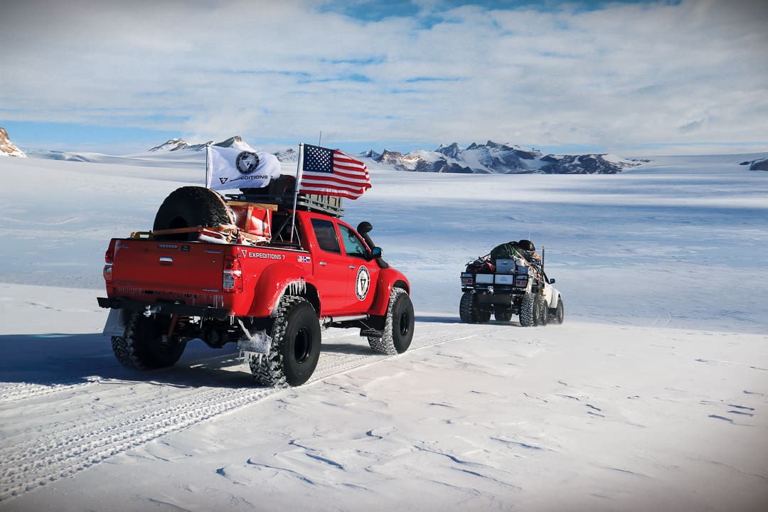 Two red expedition trucks with oversized tires drive across the vast, snow-covered landscape of the White Desert under a bright sky.