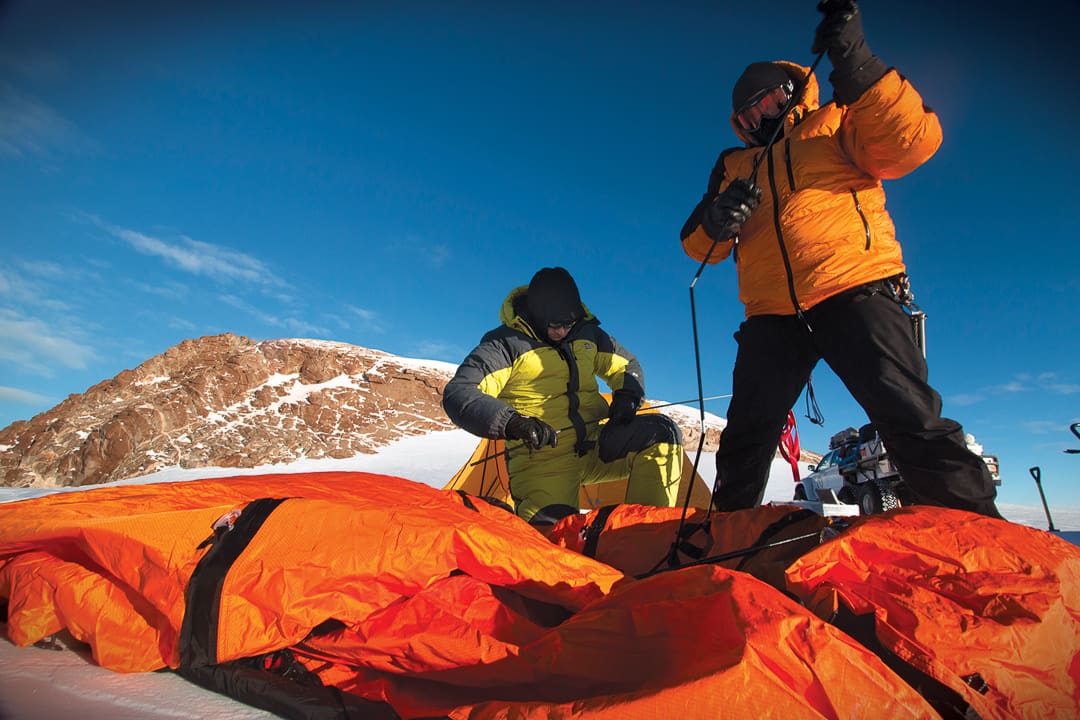 Two explorers in cold-weather gear setting up a large orange tent on a snow-covered mountain during an Antarctica expedition.