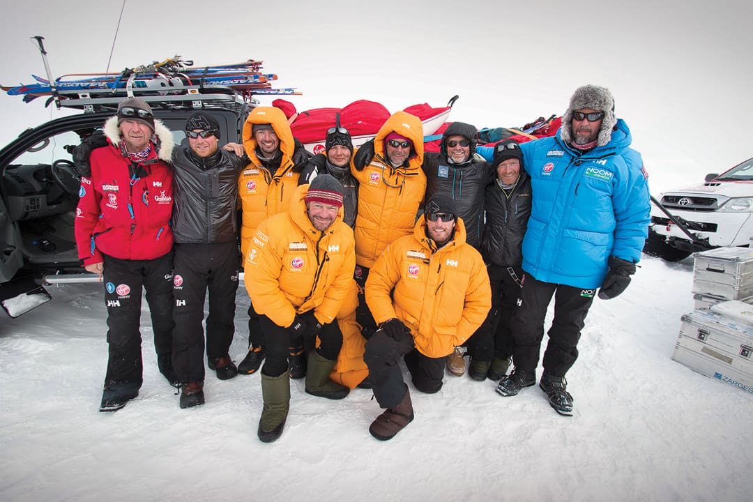 Ten people in colorful insulated expedition gear and winter hats pose on the snowy landscape of the White Desert in front of a parked vehicle with a roof rack.