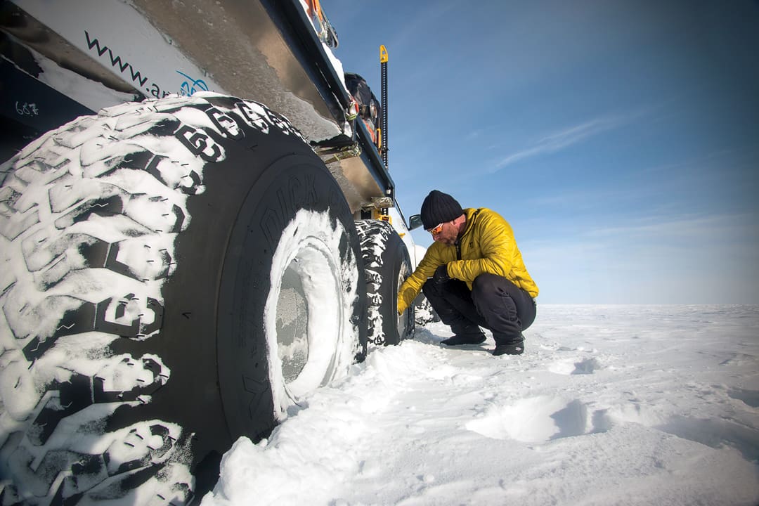 A person in a yellow jacket kneels in the snow of the White Desert to check the massive tire of an Expeditions 7 vehicle.