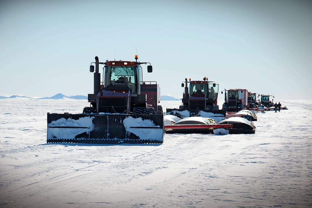 A line of tracked tractors pulling trailers across the vast, snow-covered landscape of Antarctica during Expeditions 7.