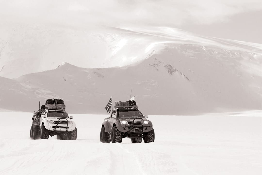 Two modified pickup trucks from Expeditions 7 with large tires drive across a vast, snow-covered landscape in the White Desert beneath snowy mountains.