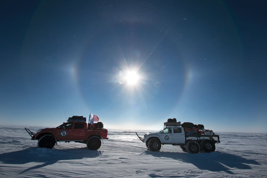 Two Expeditions 7 trucks parked on a vast, snowy White Desert under a bright sun with a sun halo.