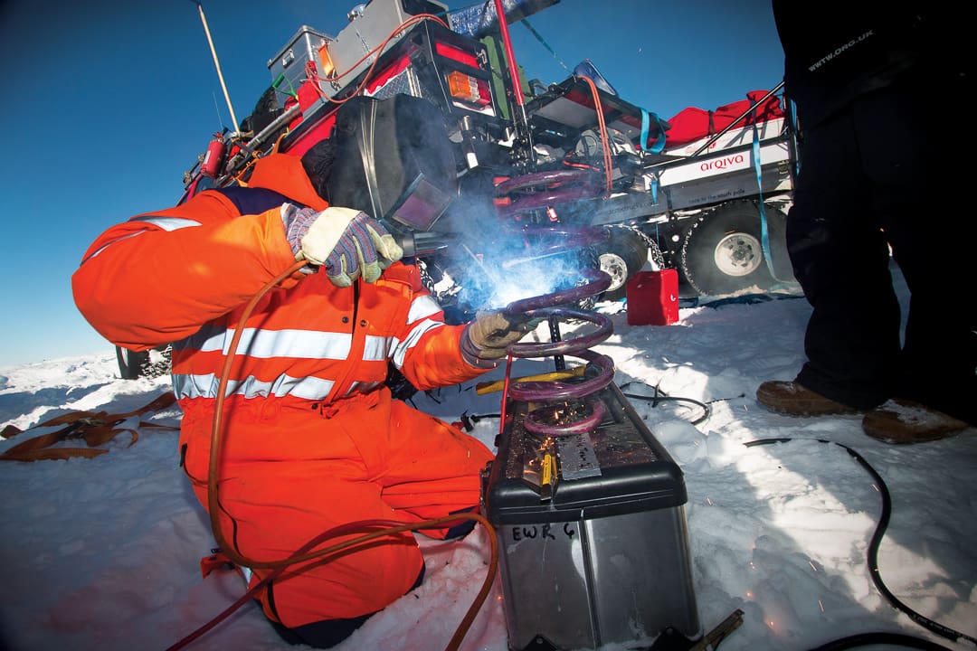 A person in orange high-visibility gear welds a metal coil in a snowy White Desert environment next to a large Expeditions 7 vehicle.