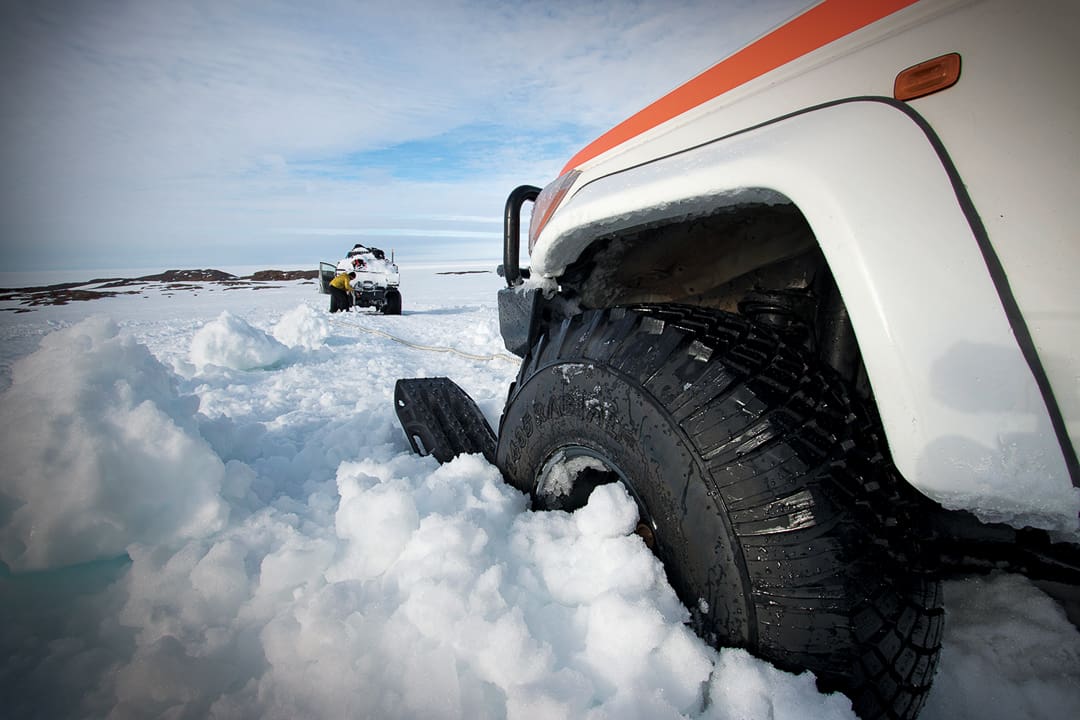 A white off-road vehicle from Expeditions 7 is stuck in deep snow while being towed by another vehicle in the distance across the white desert of Antarctica.
