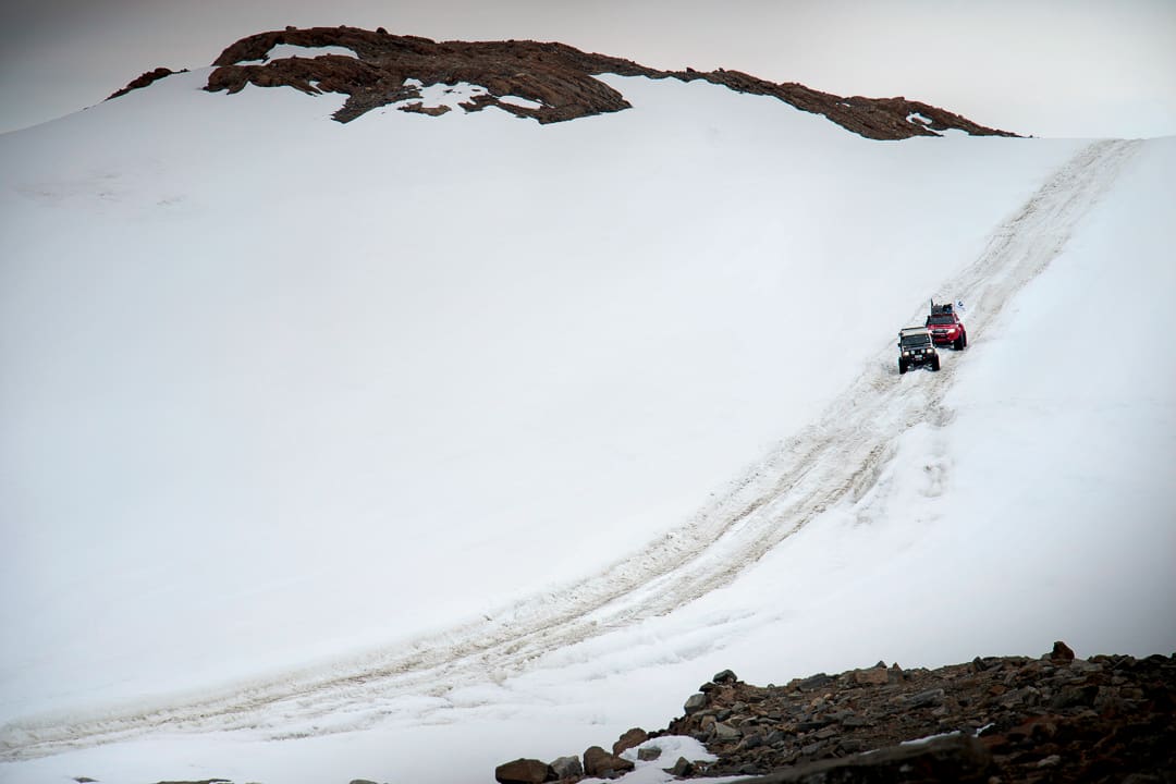 Two Expeditions 7 off-road vehicles drive up an Antarctic snowy mountain slope toward a rocky peak.