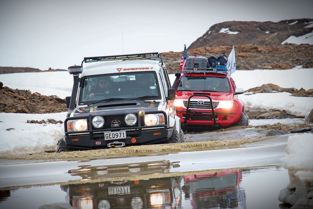 A white Land Cruiser and a red Toyota pickup truck drive through a shallow, muddy stream in the snowy, rocky landscape of Antarctica during Expeditions 7.