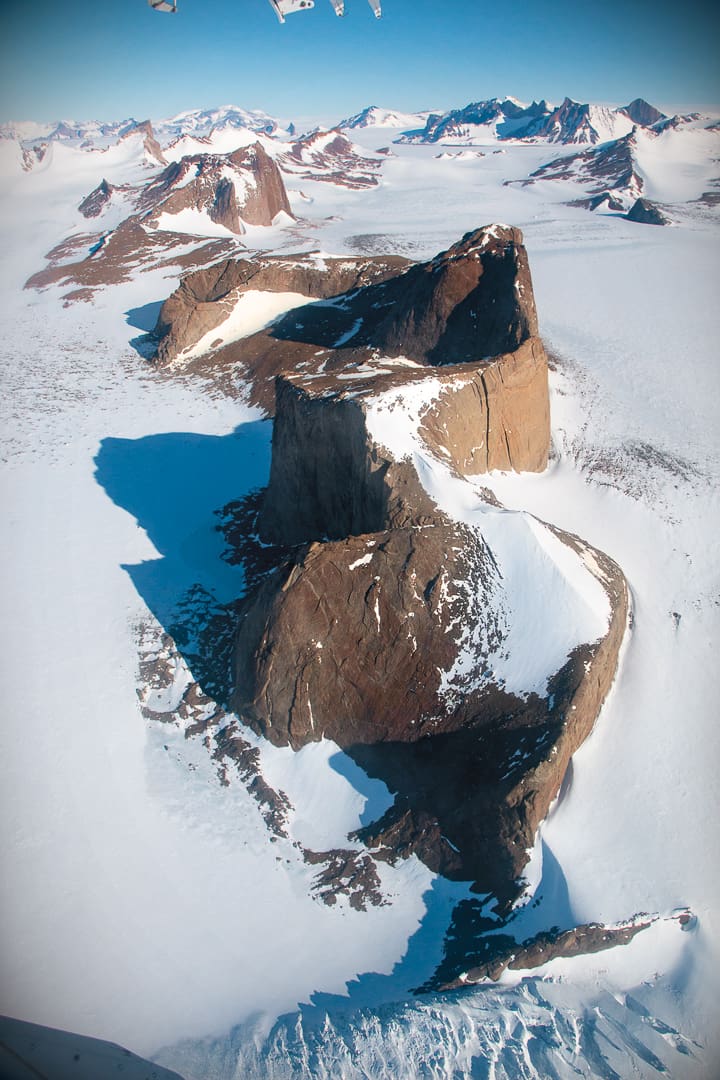 Aerial view of a jagged, dark rock formation rising out of the vast, snow-covered white desert of Antarctica.