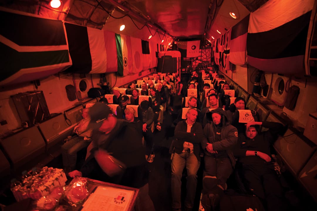 Passengers sleep in seats inside a dimly lit Expeditions 7 aircraft cabin, decorated with international flags along the walls.