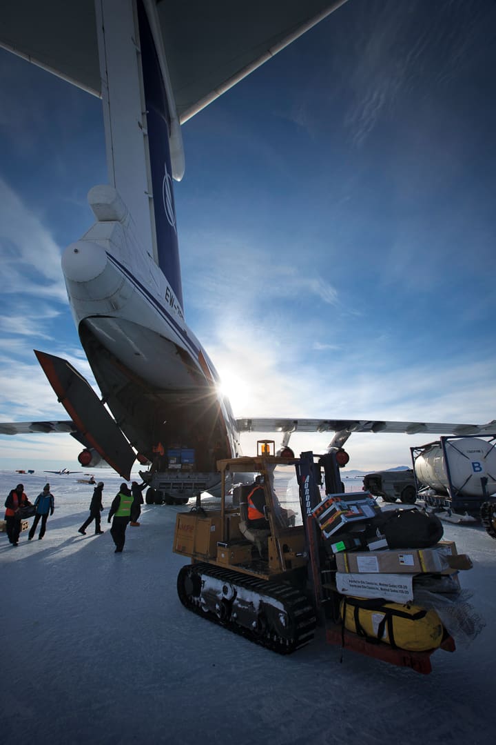 A tracked forklift carries boxes and a large bag near an open cargo plane on a snow-covered Antarctica airfield during Expeditions 7.