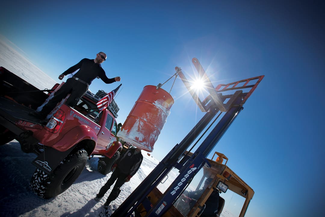 A man stands in the back of a red expedition truck as a forklift operator lowers an orange fuel drum toward him across the snow-covered Antarctica landscape.
